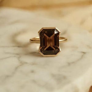 May include: A gold ring featuring a large, rectangular, smoky brown gemstone. The ring has a simple, elegant design with a thin gold band. The gemstone is set in a gold bezel, creating a clean, modern look. The ring is displayed on a white marble surface.