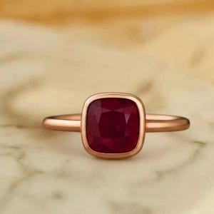 May include: A rose gold ring with a square, deep red gemstone. The ring is set on a marble surface, with the words "GARDEN MINIMALIST GARDEN" visible in the background. The ring's design is simple and elegant.