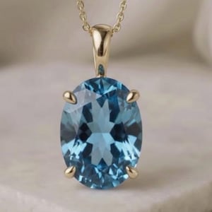 Op de afbeelding: Een gouden hangerketting met een grote, ovaal geslepen, lichtblauwe edelsteen. De edelsteen wordt op zijn plaats gehouden door vier gouden klauwen. De ketting en de hangerzetting zijn ook van goud. De ketting wordt op een wit oppervlak gepresenteerd.