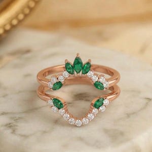 May include: Rose gold ring set with a crown of green marquise-cut gemstones and a curved band of white diamonds. The ring is on a marble surface. The text "GARDEN MINIMALIST GAR" is at the top.