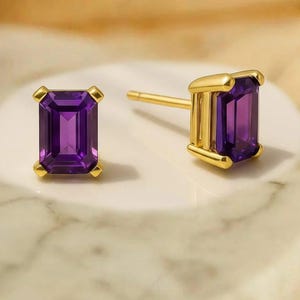 May include: A pair of gold stud earrings with rectangular, deep purple gemstones. The earrings are set in a gold frame with a post backing. The background is a marble-like surface.