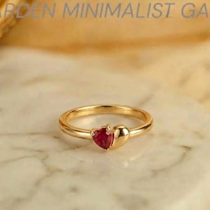 Op de afbeelding: Een gouden ring met een kleine, rode, hartvormige edelsteen naast een glad, gepolijst gouden hartvormig element. De ringband is dun en rond, gemaakt van goudkleurig metaal. De tekst GARDEN MINIMALIST GA is bovenaan in beeld zichtbaar.