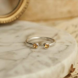 Könnte beinhalten: Ein silberner Ring mit zwei runden, hellorangen Edelsteinen an den gegenüberliegenden Enden. Der Ring ist vorne offen und hat ein minimalistisches Design. Der Ring wird auf einer Marmoroberfläche präsentiert.