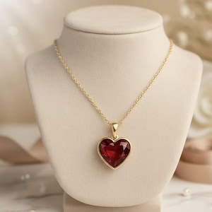 Könnte beinhalten: Eine goldfarbene Halskette mit einem herzförmigen, tiefroten Anhänger. Der Anhänger ist in einen goldenen Rahmen gefasst und hängt an einer zarten Goldkette. Die Halskette wird auf einer cremefarbenen Schmuckbüste präsentiert.