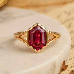 May include: A gold ring featuring a central hexagonal-cut deep red gemstone set in a bezel setting. The band has a split-shank design that connects to the sides of the hexagonal frame, creating a geometric look with polished metal surfaces.