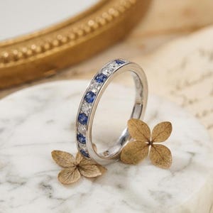 May include: A silver band ring with alternating blue sapphires and clear diamonds. The ring is set on a white marble surface, with dried flowers in the foreground. The ring is a classic design, suitable for various occasions.