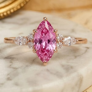 Puede incluir: Un anillo de oro rosa con una gran piedra central rosa de corte marquesa sujeta por garras. La banda está adornada a ambos lados por un grupo de pequeñas piedras redondas transparentes y una piedra de corte de pera, creando un diseño simétrico.