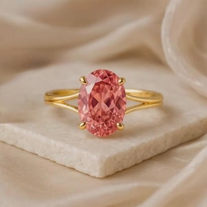 May include: A gold ring with a large, oval-cut pink gemstone. The ring has a split shank design, with the band splitting into two delicate strands on either side of the gemstone. The ring is set on a white surface.