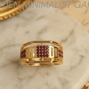 May include: A gold ring featuring a central square panel and side sections inlaid with small, round, deep red stones. The band has a multi-layered, open-work design with horizontal bars connecting the top and bottom edges of the ring structure.