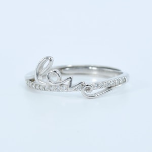 May include: A silver ring spelling out the word "love" in cursive. The ring is embellished with small, clear gemstones. The design is delicate and elegant, suitable for a romantic gift or daily wear. The ring is made of silver.