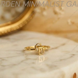 Op de afbeelding: Een gouden ring met een gepolijste metalen band en een centraal decoratief element gevormd door de in elkaar grijpende hoofdletters C en D. Het ontwerp is vervaardigd uit een glad, reflecterend goudkleurig materiaal met een afwerking.