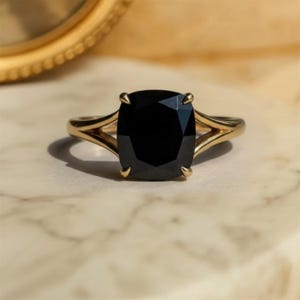 May include: A gold ring with a large, square-cut black gemstone. The ring has a split shank design, with the band splitting into two delicate curves on either side of the stone. The ring is set on a marble surface.