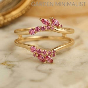 Puede incluir: Un anillo de oro con dos bandas curvas que se unen en el centro. Las bandas están adornadas con múltiples piedras preciosas rosas en cortes redondos y marquesa, dispuestas en un patrón de racimo. El texto GARDEN MINIMALIST es visible arriba.
