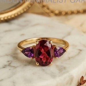 Puede incluir: Un anillo de oro con una gran gema roja oscura de corte ovalado flanqueada por dos gemas moradas triangulares más pequeñas. El anillo está sobre una superficie de mármol blanco y gris. El texto "ARDEN MINIMALIST GAR" es visible.