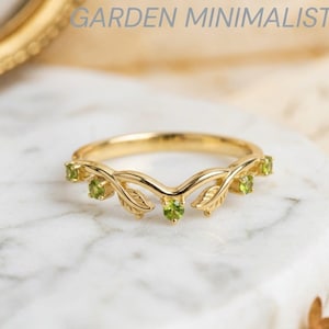 May include: A gold ring featuring a V-shaped band with engraved leaf motifs and five small round green peridot gemstones set along the top. The text GARDEN MINIMALIST is visible in the upper portion of the image above the ring.