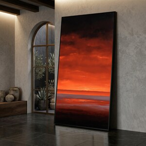 Può includere: Una grande stampa artistica incorniciata raffigurante un tramonto vibrante. L'opera presenta un gradiente di colori, che passa dal nero intenso in alto a tonalità arancioni e rosse infuocate, con accenni di bianco e viola in basso. L'opera è appoggiata a un muro grigio.