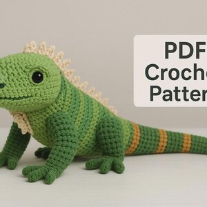 Puede incluir: Un juguete de iguana tejido a ganchillo en color verde con una cresta dorsal crema y una cola con rayas naranjas. La iguana tiene grandes ojos negros y una superficie texturizada. Un letrero muestra el texto PDF Crochet Pattern.