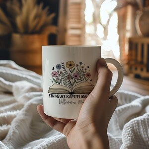 Könnte beinhalten: Weiße Keramiktasse mit floralem Design und dem Text "EIN NEUES KAPITEL BEG Willkommen 2026". Die Tasse hat einen gebogenen Henkel. Das Design umfasst Blumen, Schmetterlinge und deutschen Text.