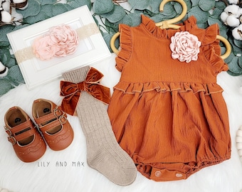 May include: A rust-colored baby onesie with a ruffled neckline and a large pink flower on the front. The onesie is paired with a pair of brown leather shoes, a pair of beige socks with a velvet bow, and a pink flower headband.