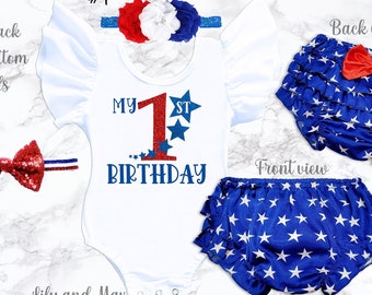 May include: A white bodysuit with red, white, and blue glitter accents, featuring the text "My 1st Birthday". The bodysuit is paired with a blue and white star-patterned diaper cover with a red bow. The image also includes two red and blue bow headbands.