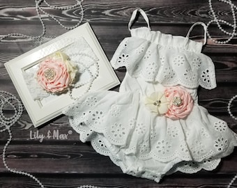 May include: White eyelet baby dress with a pink and cream flower embellishment. The dress has a ruffled neckline and a scalloped hem. The dress is paired with a matching headband with a pink and cream flower.