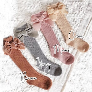 May include: Four pairs of knee-high socks in brown, gray, mauve, and cream colors, each adorned with a large velvet bow. The socks are ribbed and displayed on a white, textured surface. The colors are labeled with handwritten text.
