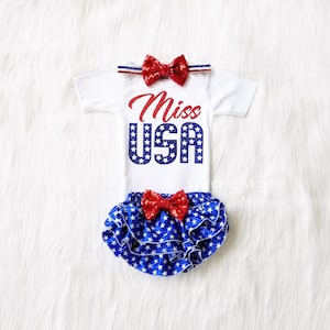 Puede incluir: Un body blanco para beb&eacute; con estrellas rojas y azules y el texto "Miss USA". El body se combina con un pa&ntilde;al de tela con estampado de estrellas azules y blancas, un lazo rojo y una diadema roja.