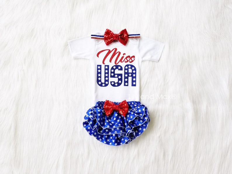 Puede incluir: Un body blanco para beb&eacute; con estrellas rojas y azules y el texto "Miss USA". El body se combina con un pa&ntilde;al de tela con estampado de estrellas azules y blancas, un lazo rojo y una diadema roja.