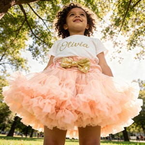 May include: A child's outfit featuring a white t-shirt with the name "Olivia" in gold script, a peach-coloured tutu skirt, and a gold sequined bow. The image is set outdoors with a background of green trees and sunlight.