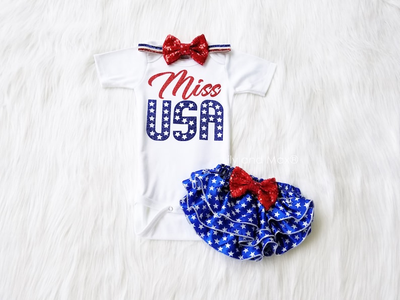 Puede incluir: Un body blanco con el texto "Miss USA" en purpurina azul y roja con estrellas blancas. Una diadema con un lazo de purpurina roja y una falda de volantes con estampado de estrellas azules y blancas con un lazo de purpurina roja.