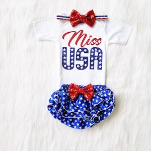 Puede incluir: Un body blanco con un dise&ntilde;o de purpurina roja y azul que dice "Miss USA". El body se combina con una cubierta de pa&ntilde;al con volantes de estampado de estrellas azul y blanco con un lazo de purpurina rojo. Una diadema con lazo de purpurina rojo completa el atuendo.
