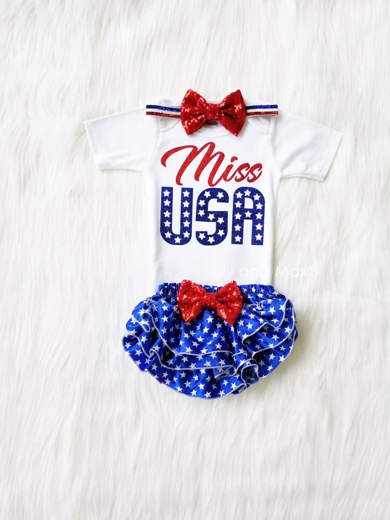 Puede incluir: Un body blanco con un dise&ntilde;o de purpurina roja y azul que dice "Miss USA". El body se combina con una cubierta de pa&ntilde;al con volantes de estampado de estrellas azul y blanco con un lazo de purpurina rojo. Una diadema con lazo de purpurina rojo completa el atuendo.