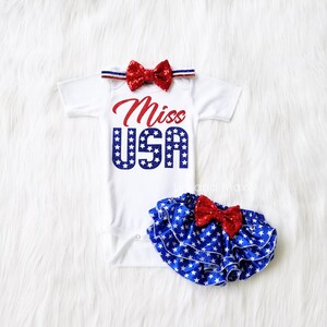 Puede incluir: Un body blanco con el texto "Miss USA" en purpurina roja y azul. El body tiene una diadema con un lazo rojo y azul y una falda de volantes de estrellas azules y blancas con un lazo rojo.