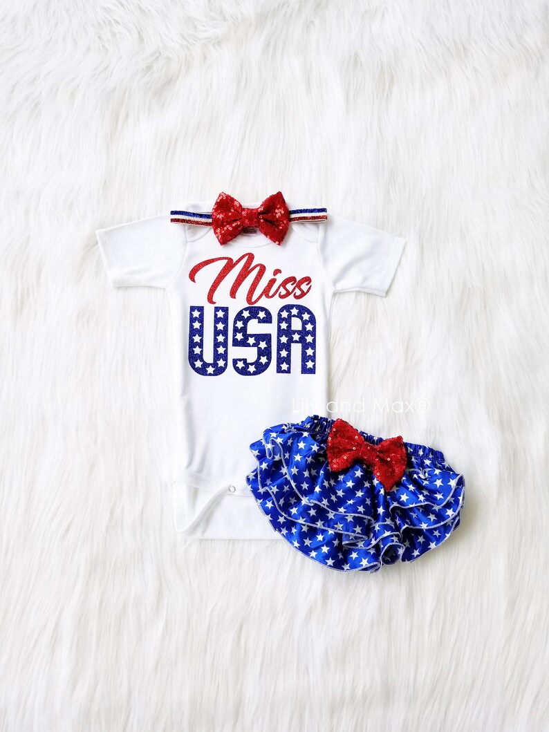 Puede incluir: Un body blanco con el texto "Miss USA" en purpurina roja y azul. El body tiene una diadema con un lazo rojo y azul y una falda de volantes de estrellas azules y blancas con un lazo rojo.