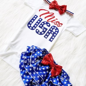 Puede incluir: Un body blanco con el texto "Miss USA" en purpurina roja y azul, combinado con una cubierta de pa&ntilde;al de volantes de estampado de estrellas azules y blancas con un lazo de lentejuelas rojas.