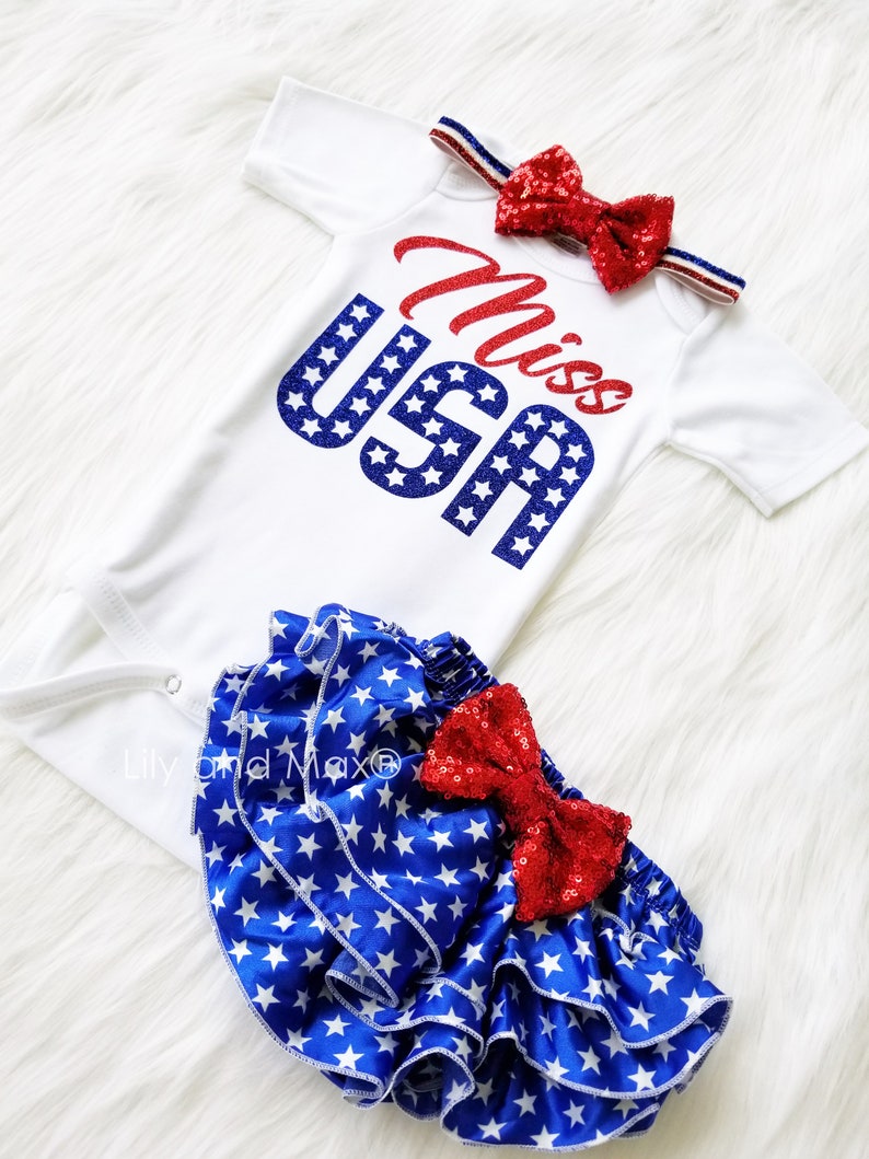 Puede incluir: Un body blanco con el texto "Miss USA" en purpurina roja y azul, combinado con una cubierta de pa&ntilde;al de volantes de estampado de estrellas azules y blancas con un lazo de lentejuelas rojas.