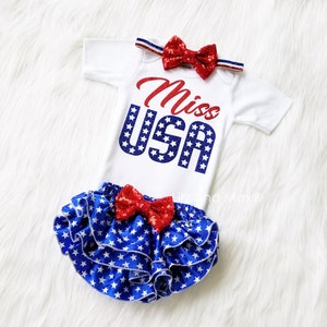 Puede incluir: Un body blanco con letras rojas y azules de purpurina que dice "Miss USA". El body se combina con una cubierta de pa&ntilde;al de volantes de estrellas azules y blancas y una diadema de lazo rojo de purpurina.
