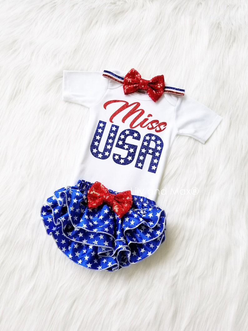 Puede incluir: Un body blanco con letras rojas y azules de purpurina que dice "Miss USA". El body se combina con una cubierta de pa&ntilde;al de volantes de estrellas azules y blancas y una diadema de lazo rojo de purpurina.