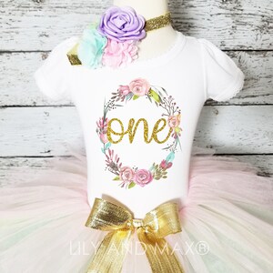 May include: A white onesie with a floral wreath and the word "one" in gold glitter. The onesie is paired with a tulle tutu in shades of pink, white, and green, and a gold bow.