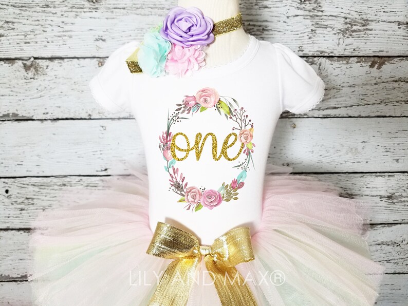 May include: A white onesie with a floral wreath and the word "one" in gold glitter. The onesie is paired with a tulle tutu in shades of pink, white, and green, and a gold bow.