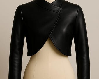 Puede incluir: Chaqueta bolero negra de manga larga con un diseño asimétrico. Destaca el cuello alto envolvente. La chaqueta se exhibe en un maniquí. El material parece ser cuero o similar al cuero.
