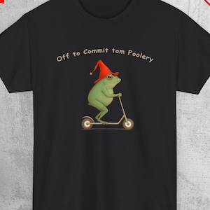 May include: Black t-shirt featuring a green frog wearing a red hat, riding a scooter. The text "Off to Commit tom Foolery" is above the frog. The shirt has promotional stickers in the corners.
