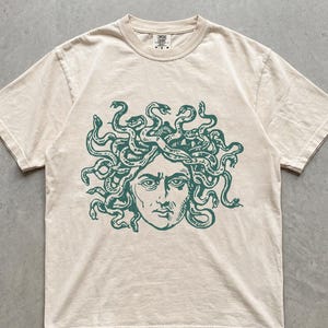 Op de afbeelding: Een ivoorkleurig t-shirt met korte mouwen en een donkergroene grafische print van een gezicht met slangen als haar op de borst. Het shirt heeft een ronde hals en een standaard pasvorm. Het woord IVORY staat linksonder in de afbeelding.