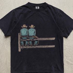 Op de afbeelding: Een zwart t-shirt met korte mouwen en een grafische illustratie van twee figuren met cowboyhoeden en blauwe overhemden die op een houten hek zitten. Het ontwerp is in het midden op de borst geplaatst en toont de figuren van achteren.