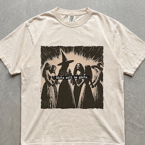 Può includere: Una t-shirt a maniche corte color avorio con una stampa grafica marrone scuro di un gruppo di streghe in cerchio. Il testo "Girls will be girls." è stampato all'interno di una barra rettangolare nera al centro dell'illustrazione.