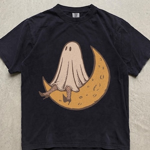 Könnte beinhalten: Ein schwarzes Kurzarm-T-Shirt mit einem Grafikdruck eines beigen Geistes, der Netzstrümpfe und hochhackige Schuhe trägt und auf einem großen gelben Halbmond sitzt. Das T-Shirt hat einen Rundhalsausschnitt und eine Standardpassform.