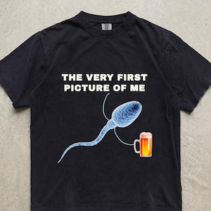 Può includere: Una t-shirt nera a maniche corte con il testo bianco THE VERY FIRST PICTURE OF ME sopra un grafico di uno spermatozoo blu che tiene in mano un boccale di birra dorata. Il disegno è centrato sul petto del capo di abbigliamento.