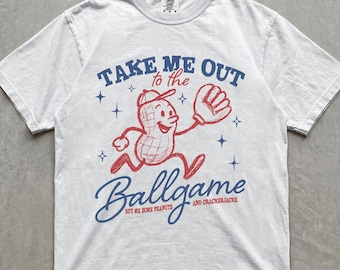 Può includere: Una t-shirt bianca a maniche corte con una grafica rossa di un personaggio a forma di arachide che indossa un berretto e un guantone da baseball. Il testo blu e rosso recita: TAKE ME OUT to the Ballgame BUY ME SOME PEANUTS AND CRACKERJACKS, con stelle.