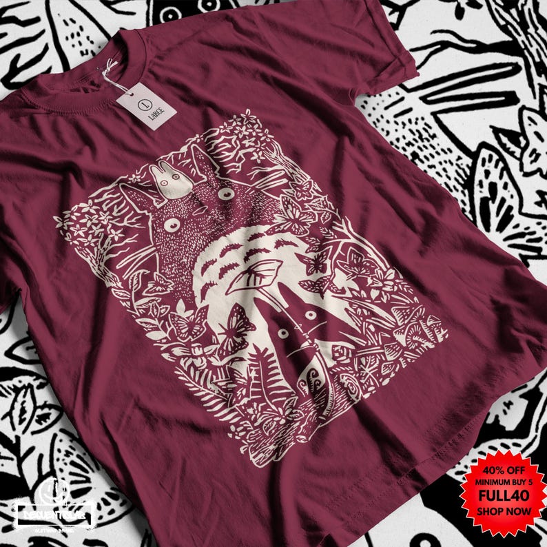 Pu&ograve; includere: T-shirt bordeaux con grafica color crema di una scena forestale con due personaggi dei cartoni animati. Il design include farfalle e fogliame. Un'etichetta &egrave; attaccata alla maglietta. Testo promozionale nell'angolo in basso a destra.