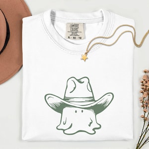 Könnte beinhalten: Ein weißes T-Shirt mit einer grünen Grafik eines Geistes, der einen Cowboyhut trägt. Das T-Shirt hat ein Comfort Colors Markenetikett am Kragen und wird zusammen mit einem beigen Hut, einer goldenen Sternkette und Trockenblumen gezeigt.